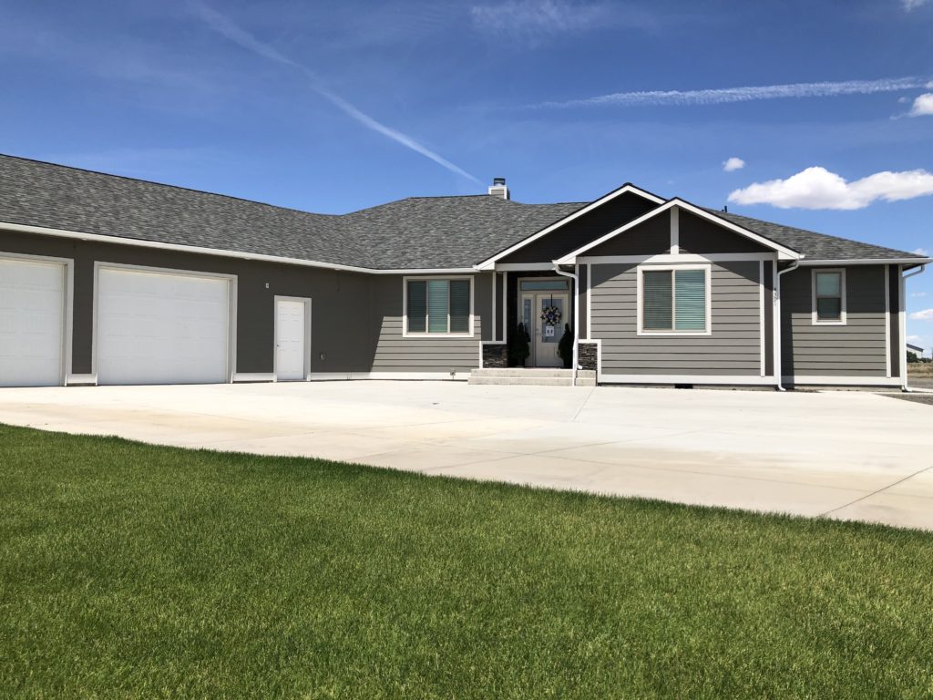 Modern gray ranch-style house with a three-car garage and a manicured lawn under a partly cloudy blue sky.